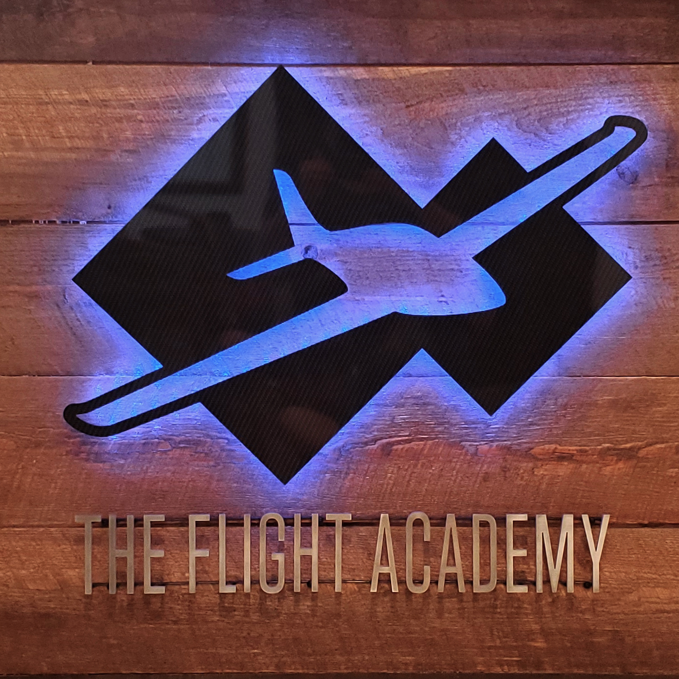 Signage for The Flight Academy, featuring their airplane logo against a wood panel, backlit in blue LED, with the name in aluminum lettering underneath.