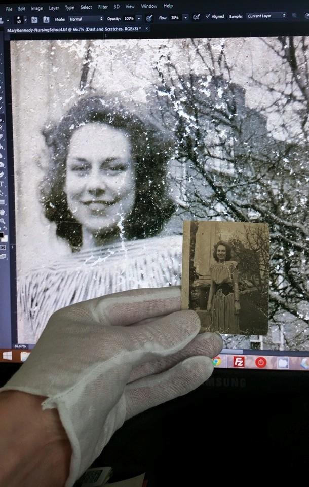 A computer monitor showing a damaged black and white image of a smiling woman being worked in Photoshop, in front of the monitor is a hand in a cotton glove, holding the original photograph.